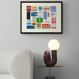 May include: A framed print of a collection of vintage matchbox labels, featuring various designs and text, including "Place", "Cocktails", "South", "Palma", "Seafarer", and "Chick's". The print is displayed on a white wall above a white sofa and a side table with a unique lamp.