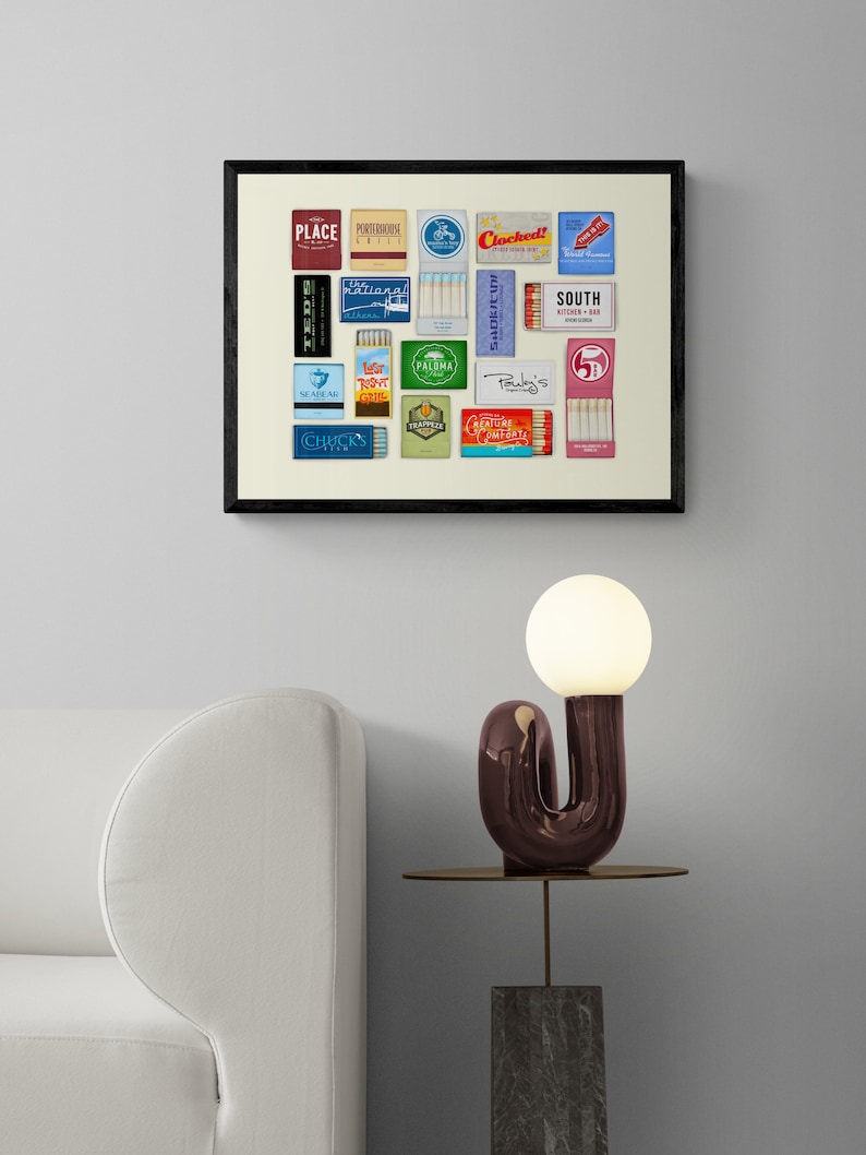 May include: A framed print of a collection of vintage matchbox labels, featuring various designs and text, including "Place", "Cocktails", "South", "Palma", "Seafarer", and "Chick's". The print is displayed on a white wall above a white sofa and a side table with a unique lamp.