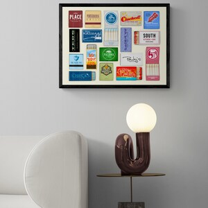 May include: A framed collage of vintage matchbook covers featuring various restaurant and bar names, including "Place", "Portaroux Grill", "Clocked", "National", "Seabear", "Lesr Roast Grill", "Paloma", "Pauley's", "Creature Comfort", "South Kitchen & Bar", and "Chuck's Fish". The collage is displayed on a white wall above a white couch and a side table with a modern, abstract lamp.