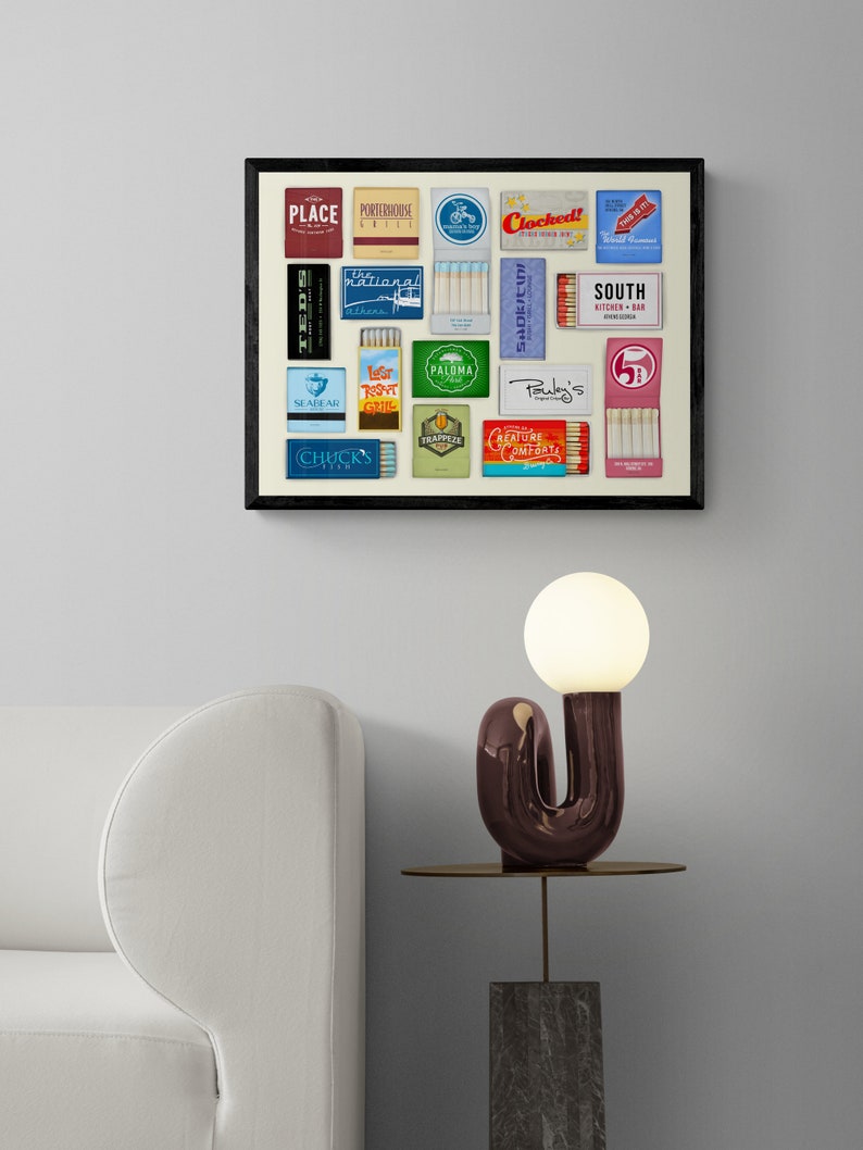 May include: A framed collage of vintage matchbook covers featuring various restaurant and bar names, including "Place", "Portaroux Grill", "Clocked", "National", "Seabear", "Lesr Roast Grill", "Paloma", "Pauley's", "Creature Comfort", "South Kitchen & Bar", and "Chuck's Fish". The collage is displayed on a white wall above a white couch and a side table with a modern, abstract lamp.