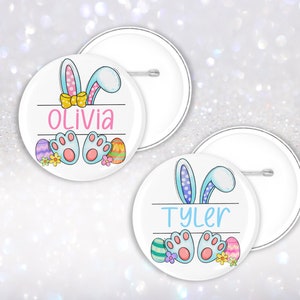 May include: Two round white buttons featuring a blue bunny ear design and colourful Easter eggs. The buttons are personalised with the names Olivia and Tyler.