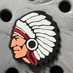 May include: A shoe charm featuring a Native American headdress design. The charm has a black background with a white feathered headdress, a red headband, and a profile of a face in shades of peach and brown. The charm is round with a black circle in the center.