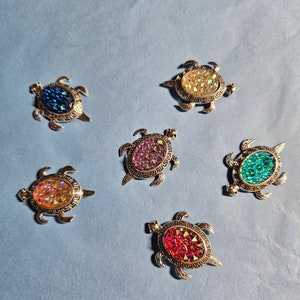 May include: Six turtle-shaped brooches with iridescent glass accents. The brooches are silver-toned and feature a variety of colors, including blue, green, pink, and yellow.