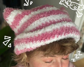 Könnte beinhalten: Eine rosa-weiß gestreifte Katzenohrmütze. Die Mütze besteht aus einem weichen, flauschigen Material und hat zwei spitze Ohren oben. Die Mütze wird von einer Person mit hellbraunem Haar und geschlossenen Augen getragen.