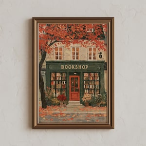 May include: A framed wall art print depicting a bookshop storefront with a dark green facade, a bright red door, and large windows filled with books. The scene features autumn foliage in shades of orange and red, with fallen leaves on the ground.
