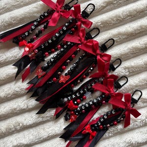 May include: Several keychains with black and red ribbons, black beads, and red bows. Each keychain has a silver clasp and a name spelled out in white letter beads. The names include: SEOKJIN, JUNGKOOK, NAMJOON, TAEHYUNG, YOONGI, and JHOPE.