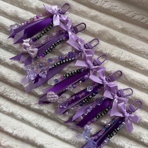 May include: A collection of purple keychains with beaded accents, ribbons, and bows. Each keychain features a name spelled out in black letter beads. The keychains are attached to a silver-colored clasp.