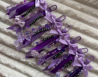May include: A collection of purple keychains with beaded accents, ribbons, and bows. Each keychain features a name spelled out in black letter beads. The keychains are attached to a silver-colored clasp.