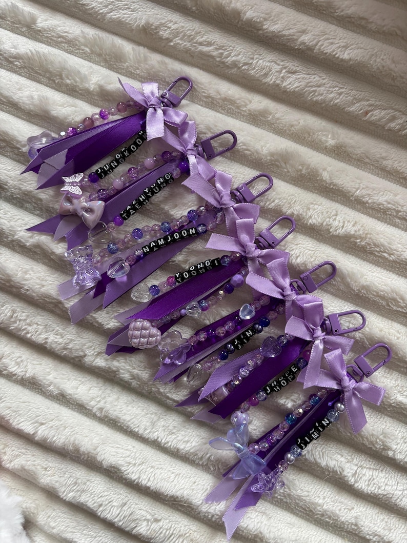 May include: A collection of purple keychains with beaded accents, ribbons, and bows. Each keychain features a name spelled out in black letter beads. The keychains are attached to a silver-colored clasp.