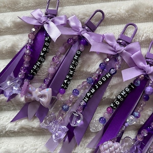 May include: Purple keychains with beaded accents, ribbons, and bows. Each keychain features a name spelled out with black letter beads. The keychains have a lobster clasp for easy attachment. The keychains are a mix of purple shades.