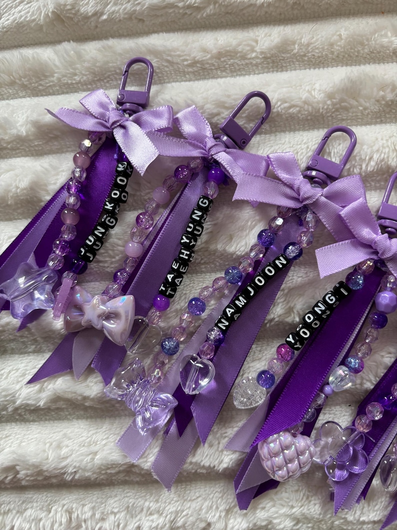 May include: Purple keychains with beaded accents, ribbons, and bows. Each keychain features a name spelled out with black letter beads. The keychains have a lobster clasp for easy attachment. The keychains are a mix of purple shades.