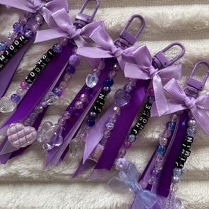 May include: Purple keychains with beaded accents, ribbons, and bows. Each keychain features a name spelled out in black letter beads. The keychains have a variety of clear and purple beads, and heart-shaped charms.