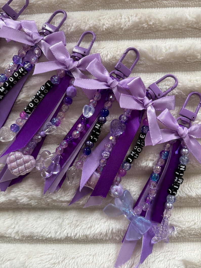 May include: Purple keychains with beaded accents, ribbons, and bows. Each keychain features a name spelled out in black letter beads. The keychains have a variety of clear and purple beads, and heart-shaped charms.