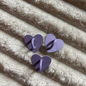 May include: Three heart-shaped wall hooks in shades of purple. Each hook has a curved, matching-color handle extending from the center. The hooks are arranged on a textured, white surface.