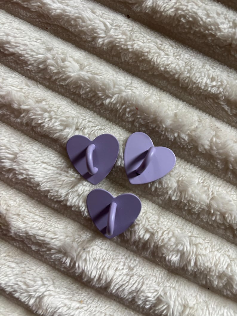 May include: Three heart-shaped wall hooks in shades of purple. Each hook has a curved, matching-color handle extending from the center. The hooks are arranged on a textured, white surface.