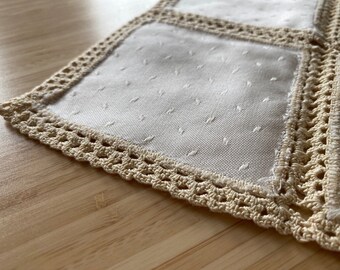 May include: Close-up of a cream-colored table runner with a crocheted border. The runner is made of a textured fabric with small, evenly spaced dots. The crocheted border is a light beige color.
