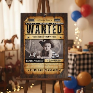 Pode incluir: Um cavalete de madeira exibe um pôster de "Procurado" para uma festa de aniversário. O pôster apresenta uma foto em tons de sépia de um bebê com um chapéu de cowboy, o texto "PROCURADO" e o nome "Daniel Gallego". O pôster inclui o texto "The Birthday Boy" e "$10.000 de recompensa".