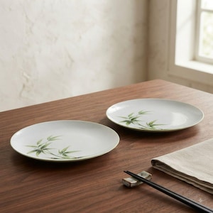 May include: Two round white ceramic plates featuring a delicate green bamboo leaf illustration. The plates are positioned on a dark wood surface alongside a pair of black chopsticks resting on a small rectangular ceramic holder and a folded cloth napkin.