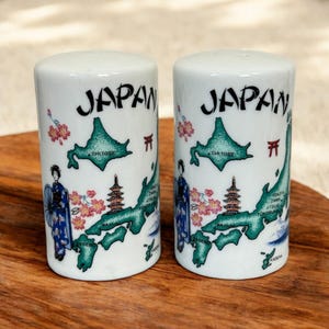 May include: Two white ceramic salt and pepper shakers with a Japan theme. Each shaker features a map of Japan, the word "JAPAN", a geisha, cherry blossoms, and a pagoda. The shakers are cylindrical and have a glossy finish.