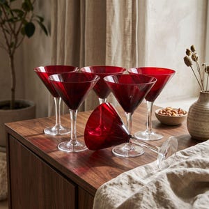May include: A set of five martini glasses with deep red bowls and clear glass stems and bases. Four glasses stand upright in a cluster, while one glass lies on its side in the foreground. The glasses feature a classic conical shape.