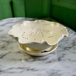 May include: Off-white ceramic colander with a wavy rim and leaf-like embossed details. The colander has several small holes and sits atop a matching saucer. The design is simple and elegant, suitable for kitchen use.