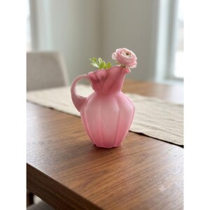 May include: A small, frosted pink glass pitcher with a scalloped, ribbed body and a curved handle. A single, light pink ranunculus flower with green leaves is placed inside the pitcher, extending from the top opening of the vessel.