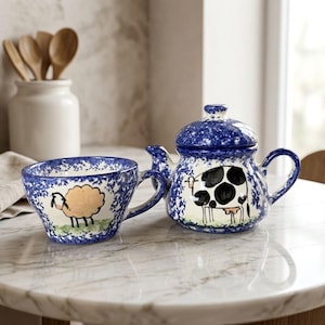 May include: A ceramic teapot and matching mug featuring a blue and white speckled pattern. The teapot displays a black and white cow illustration, while the mug features a beige sheep illustration. Both pieces have a rounded shape and handles.