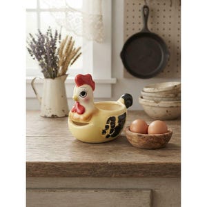 May include: A ceramic chicken-shaped egg holder with a cream-coloured body, a bright red comb and wattle, and black painted wing details. The chicken has large eyes with long eyelashes and is positioned next to a small wooden bowl containing two brown eggs.