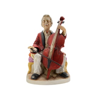 Puede incluir: Figura de cerámica de un anciano tocando el violonchelo. El hombre está sentado, con una chaqueta roja, pantalones blancos y zapatos negros. El violonchelo es de color rojo intenso y la figura tiene un aspecto detallado y realista.