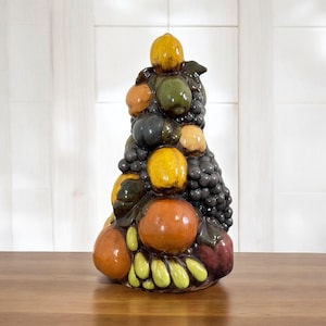 May include: A ceramic fruit sculpture featuring a variety of colorful fruits. The sculpture includes yellow lemons, orange oranges, green pears, and dark purple grapes. The fruits are arranged in a stacked, conical shape, creating a decorative piece.