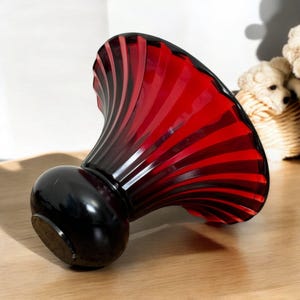 May include: A decorative glass perfume bottle with a black base and a red, fan-shaped top. The bottle is lying on its side on a wooden surface. The top has a ribbed design, and the bottle has a glossy finish.
