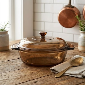 May include: A round, amber-colored glass casserole dish with a matching lid and two side handles. The transparent cookware features a smooth, polished finish and a central knob on the lid, designed for heat-resistant use in a kitchen setting.