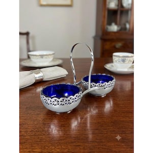 May include: A silver-toned metal condiment server featuring two round bowls with deep blue glass inserts. The bowls are encased in a decorative filigree lattice frame and connected by a central arched handle for carrying.