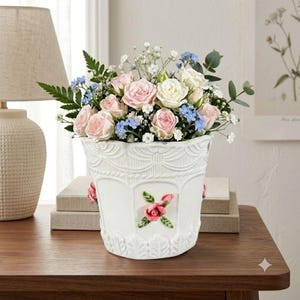 May include: A white ceramic vase with embossed swags and a central floral relief featuring a pink rose. The vase is filled with a bouquet of light pink roses, white roses, small blue flowers, baby's breath, and green eucalyptus leaves.