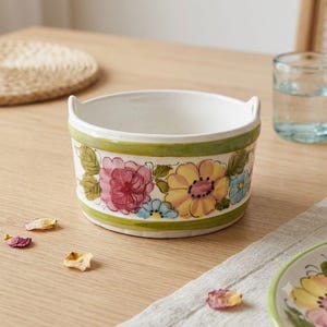 May include: A round ceramic bowl with two small side handles, featuring a white interior and a hand-painted exterior band of green stripes framing a colorful floral pattern of pink, yellow, and blue flowers with green leaves on a white base.