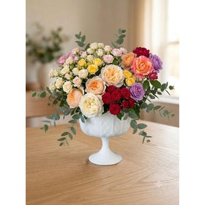 Peut inclure: Un arrangement floral vibrant composé de roses pêche, jaunes, rouges et violettes avec des feuilles d'eucalyptus vertes, présenté dans un bol sur pied blanc orné d'un motif de feuilles texturé sur la surface extérieure du récipient.