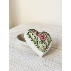 Puede incluir: Un joyero de cerámica en forma de corazón con una base de color crema. La tapa presenta un diseño en relieve pintado a mano de rosas rosadas rodeadas de intrincadas enredaderas verdes y patrones de follaje, con un acabado liso y brillante.