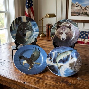 May include: Four round decorative ceramic plates displayed on black stands. One plate features a gray wolf, one a brown bear, one a bald eagle in flight, and one a salmon jumping over a waterfall. Each plate displays detailed wildlife imagery.