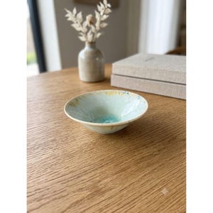 May include: A shallow ceramic bowl with a wide rim, featuring a reactive glaze in shades of pale blue, cream, and hints of yellow. The interior base has a concentrated pool of deep turquoise color, while the exterior surface is smooth.