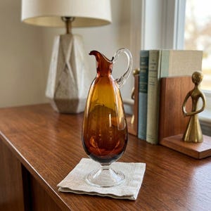 May include: A teardrop-shaped amber glass pitcher with a clear glass handle and a clear glass pedestal base. The pitcher features a narrow neck and a small pouring spout, showcasing a smooth, translucent finish throughout the glass body.