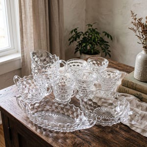 May include: A collection of clear pressed glass tableware featuring a repeating geometric diamond pattern. The set includes a pitcher, several handled cups, a long oval serving dish, and a round bowl, all with a textured, faceted surface.