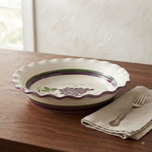 May include: A round ceramic pie dish with a ruffled edge, featuring a cream-colored interior decorated with a painted cluster of purple grapes and green leaves, accented by a dark purple and green band around the inner rim.