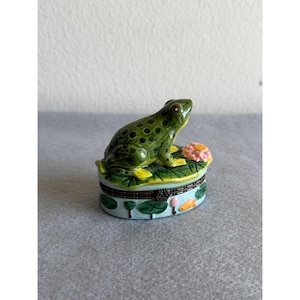 Pode incluir: Caixa de bugigangas decorativa com um sapo verde em cima de uma folha de nenúfar com uma flor rosa. A caixa tem uma base azul clara com nenúfares e peixes pintados. A tampa articulada tem uma moldura de metal.