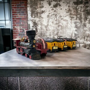 Könnte beinhalten: Eine rote und schwarze Spielzeugeisenbahn mit drei gelben Spielzeugwaggons. Die Lokomotive hat einen schwarzen Schornstein und einen roten Schlusswagen. Die Waggons haben schwarze Räder und Fenster. Die Eisenbahn steht auf einer grauen Oberfläche.