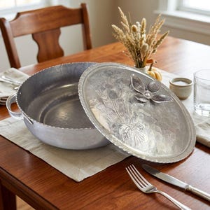 May include: A round, hammered aluminum casserole dish with two side handles and a matching lid. The lid features an embossed floral bouquet design and a three-dimensional metal flower handle. The metal surface has a textured, matte finish.
