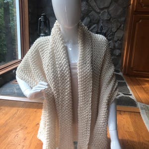 May include: Cream-colored, hand-knitted shawl draped over a mannequin. The shawl features a textured stitch pattern and a draped design. The neutral color and cozy texture suggest a focus on comfort and warmth. The shawl is displayed on a mannequin with a neutral background.