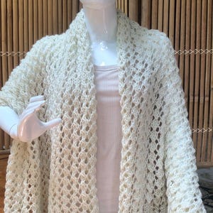 Puede incluir: Un chal de ganchillo de color blanco roto con un patrón de punto abierto en forma de rombos. La prenda está colocada sobre un maniquí blanco, mostrando la intrincada y aireada textura del hilo y los bordes festoneados del dobladillo.