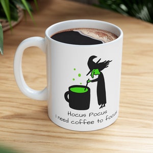Op de afbeelding: Witte keramische koffiemok met een zwart-groene illustratie van een heks die een ketel roert. De tekst "Hocus Pocus I need coffee to focus" staat op de mok gedrukt.