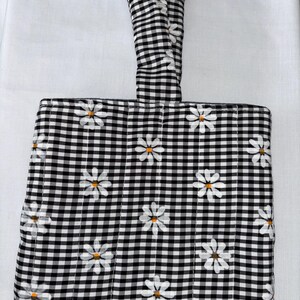 May include: A quilted black and white gingham cutting board cover with a handle. The cover is decorated with white daisies with yellow centres. The handle is in a V-shape. The cover is on a white surface.