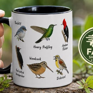 May include: A white ceramic mug with a black interior and handle, featuring colorful illustrations of six different birds labeled Bushtit, Hoary Puffleg, Andean Flicker, Red-billed Woodpecker, Woodcock, and Dickcissel printed on the exterior surface.